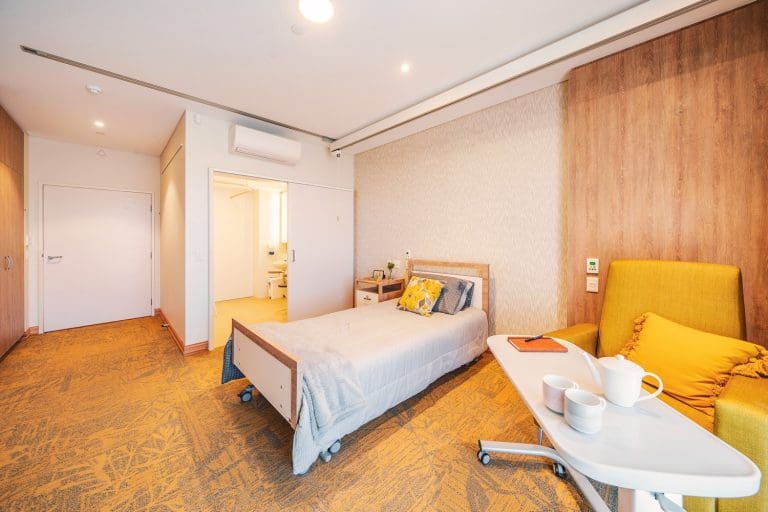 Aged care suite with comfortable chair, bed and private ensuite bathroom in the background